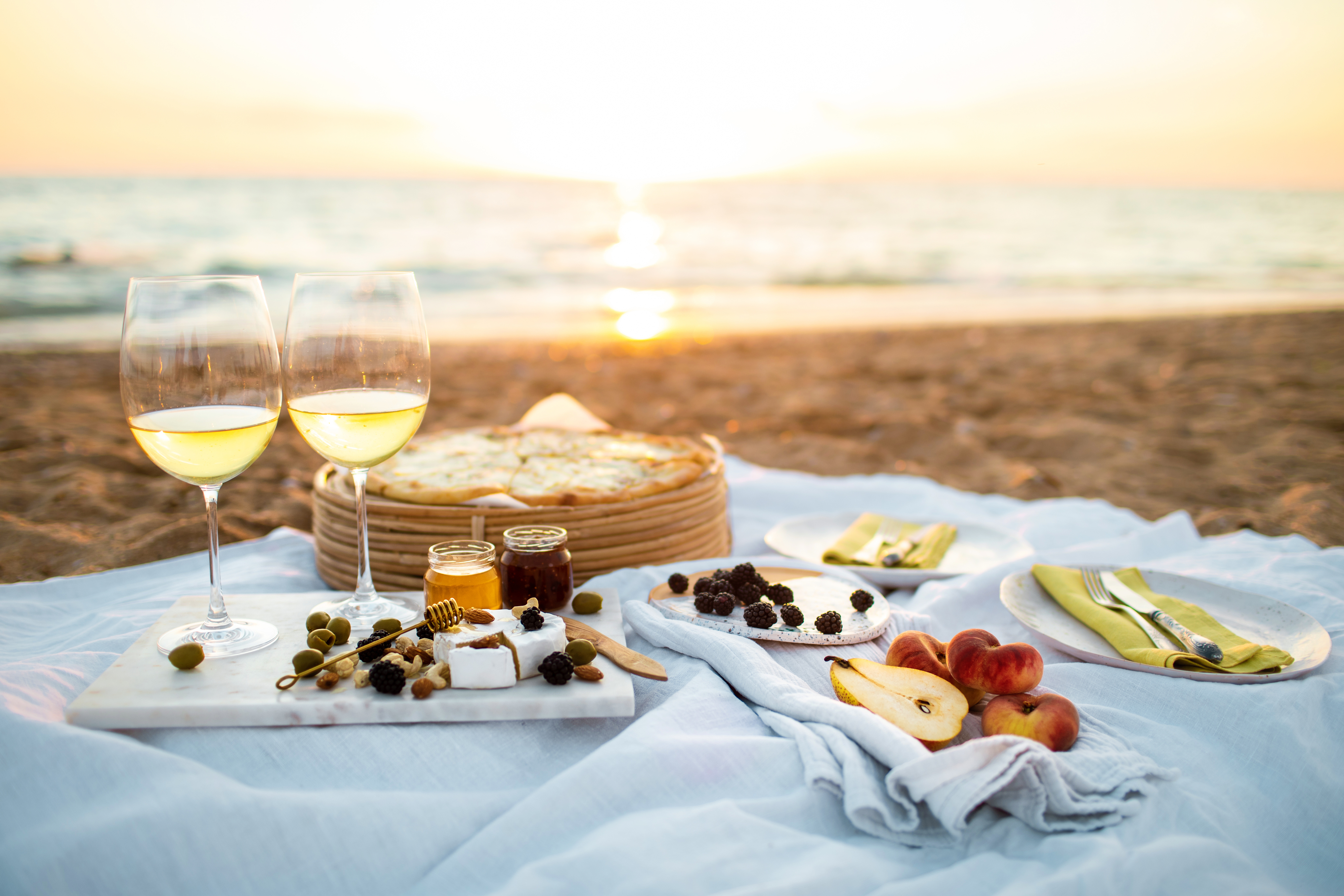 A beach picnic at sunset featuring wine glasses, cheese, fruit, nuts, and bread laid out on a white blanket by the shore