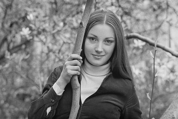 A woman with long hair, wearing a collared shirt and jacket, stands outdoors holding a tree branch