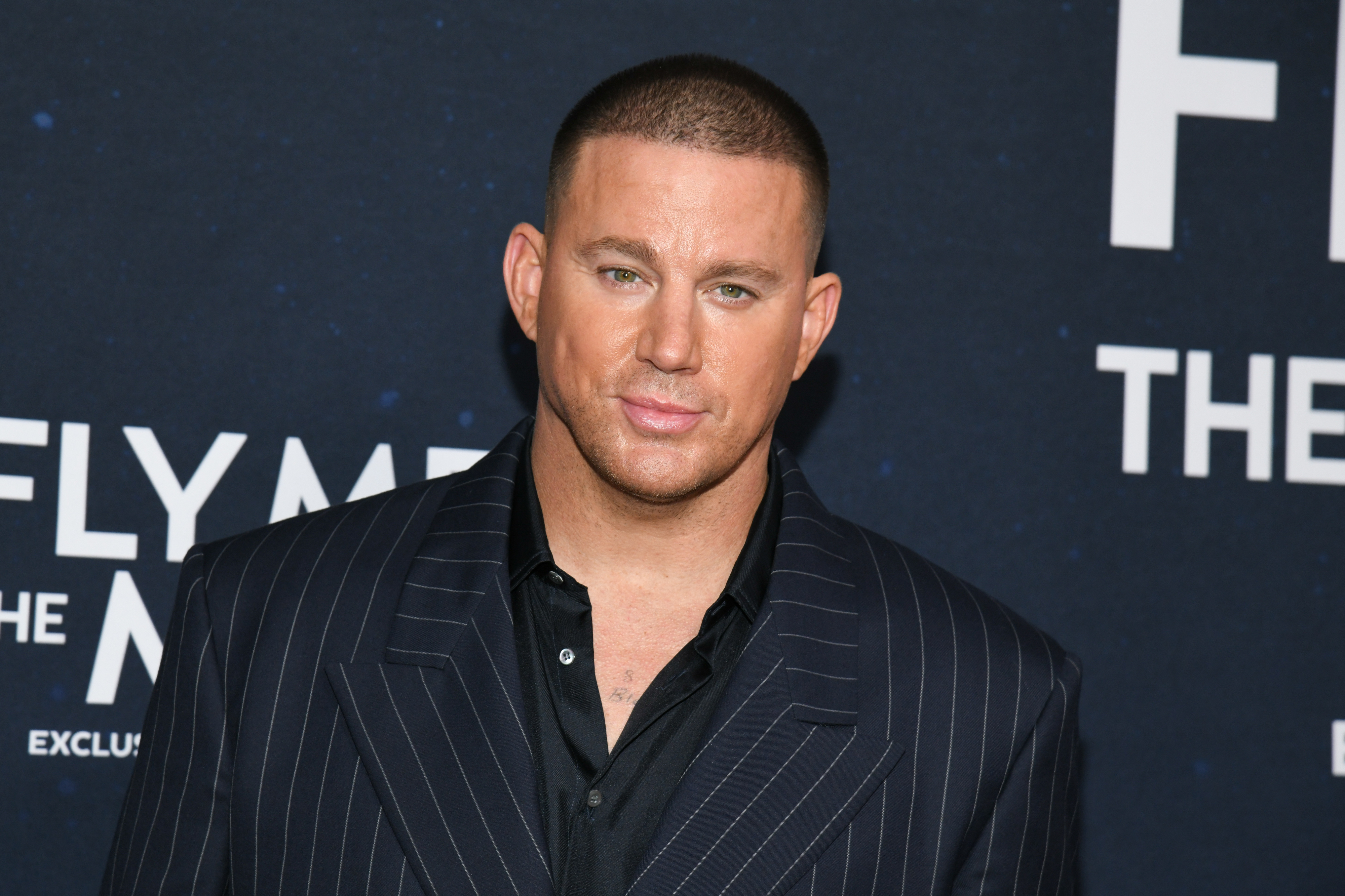 Channing Tatum at an event, wearing a dark pinstripe suit with an open-collared shirt, standing in front of a backdrop that reads "Fly Me to the Moon"