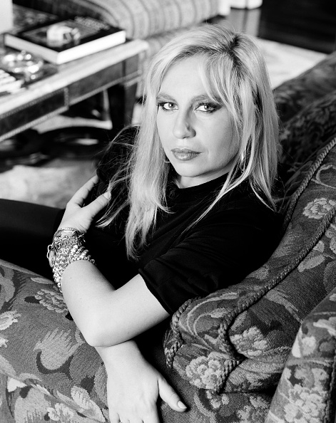 A black and white image of a woman with long blonde hair, wearing a black top and bracelets, sitting on a sofa and looking at the camera