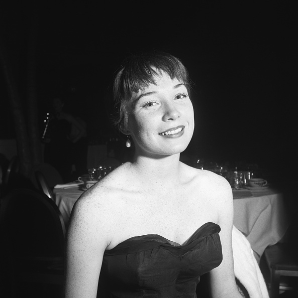 A woman in a strapless dress smiles at the camera. The background features a table with glassware and another person
