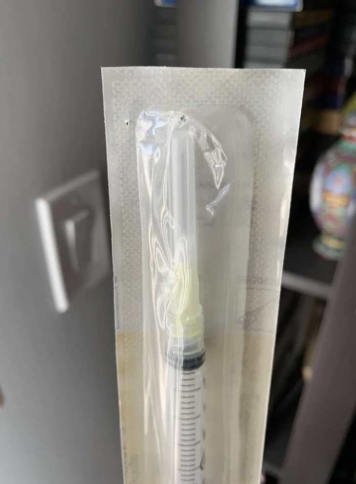 Sterile vacuum-sealed medical syringe in packaging, standing upright against a background that includes part of a bookshelf and a decorative vase
