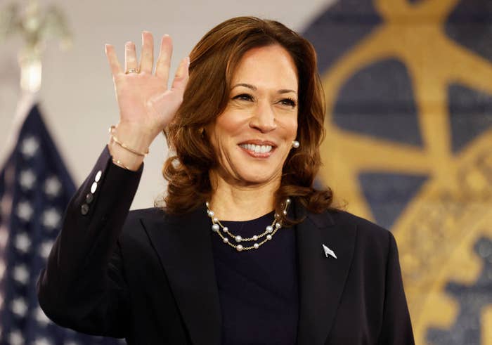 Kamala Harris waves and smiles while wearing a dark blazer, a pearl necklace, and an American flag pin in front of U.S. flags and a rotary emblem