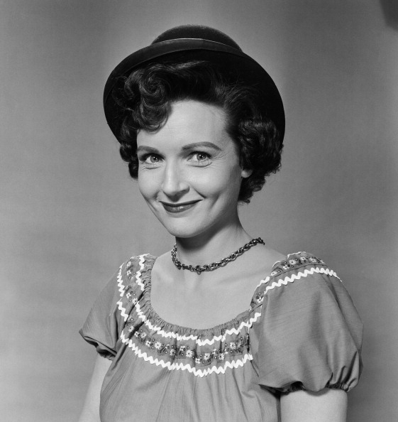 Black and white photo of older celeb wearing a hat, a short-sleeved dress with decorative embroidery, and a chain necklace