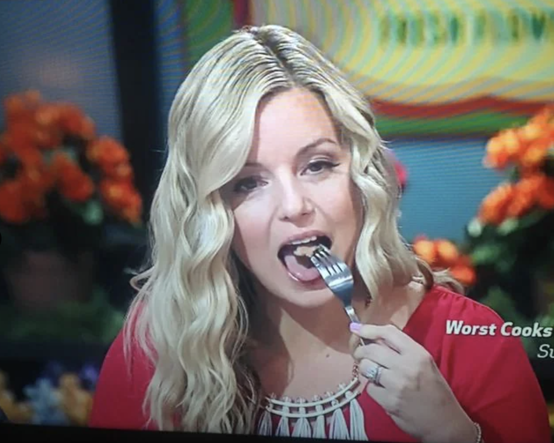 A woman with long, wavy hair is eating from a fork on the set of "Worst Cooks in America." Flowers are in the background. Text on the screen reads "Worst Cooks in America."