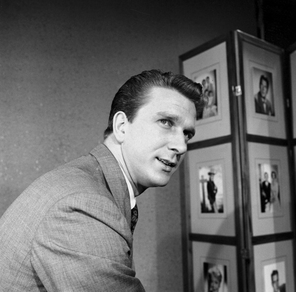 A man in a suit poses for a photograph; framed photos are visible in the background