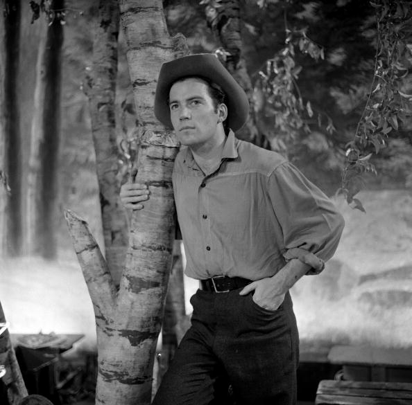 Man in a cowboy hat and casual shirt leaning against a tree in a forest setting