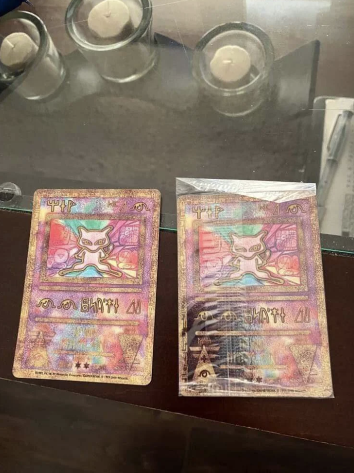 Two collectible cards featuring the character Mew from Pokémon are displayed side by side on a table with candles and a pen in the background