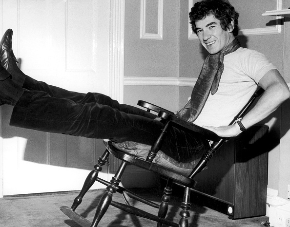 A person is smiling and leaning back with legs extended in a rocking chair, they are dressed in a t-shirt, scarf, and pants