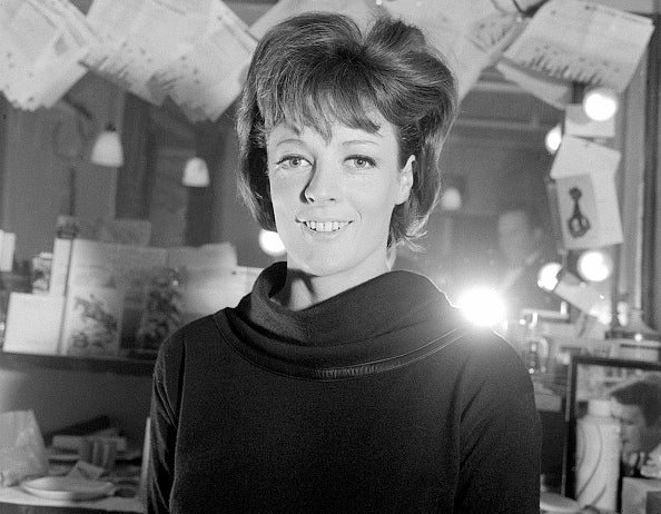 A woman with an elaborate hairstyle, wearing a dark top, stands smiling in a room with papers and lights hanging from the ceiling