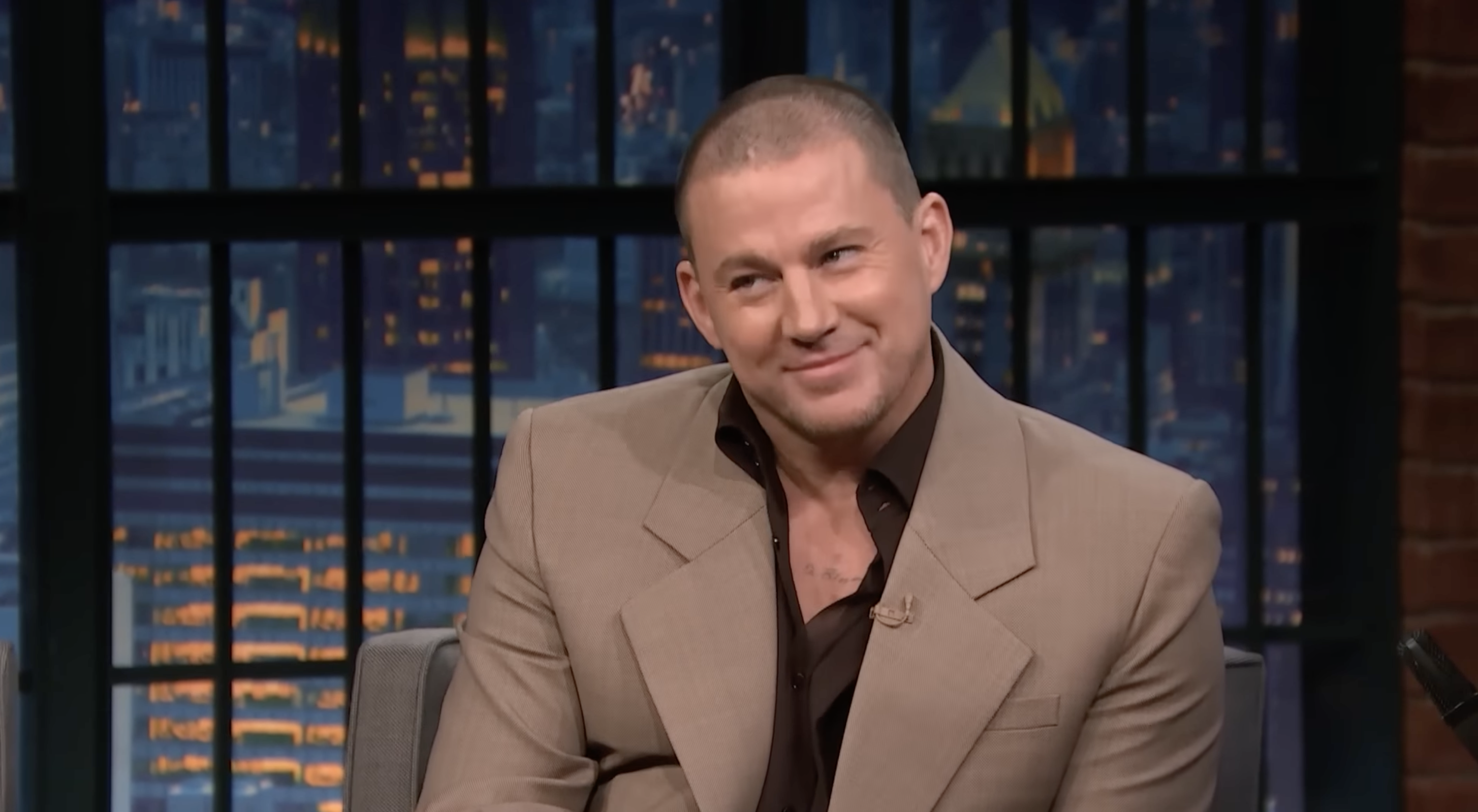 Channing Tatum smiling during a talk show appearance, wearing a beige suit and a dark shirt. Cityscape is visible through the window behind him