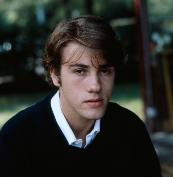 A person with medium-length hair is wearing a collared shirt under a sweater and looking directly at the camera outdoors
