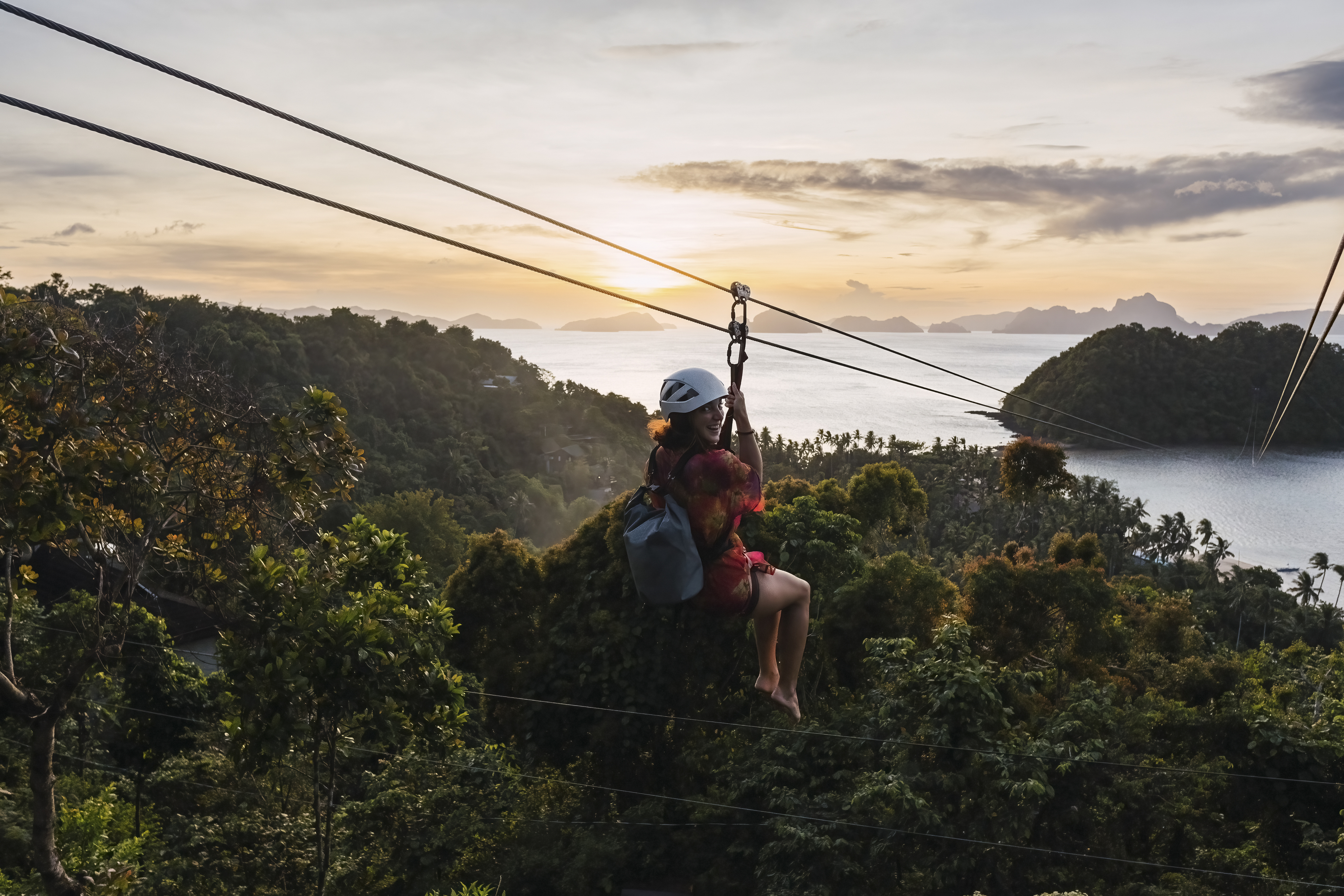 Person ziplining through a lush tropical landscape with mountains and ocean in view during sunset