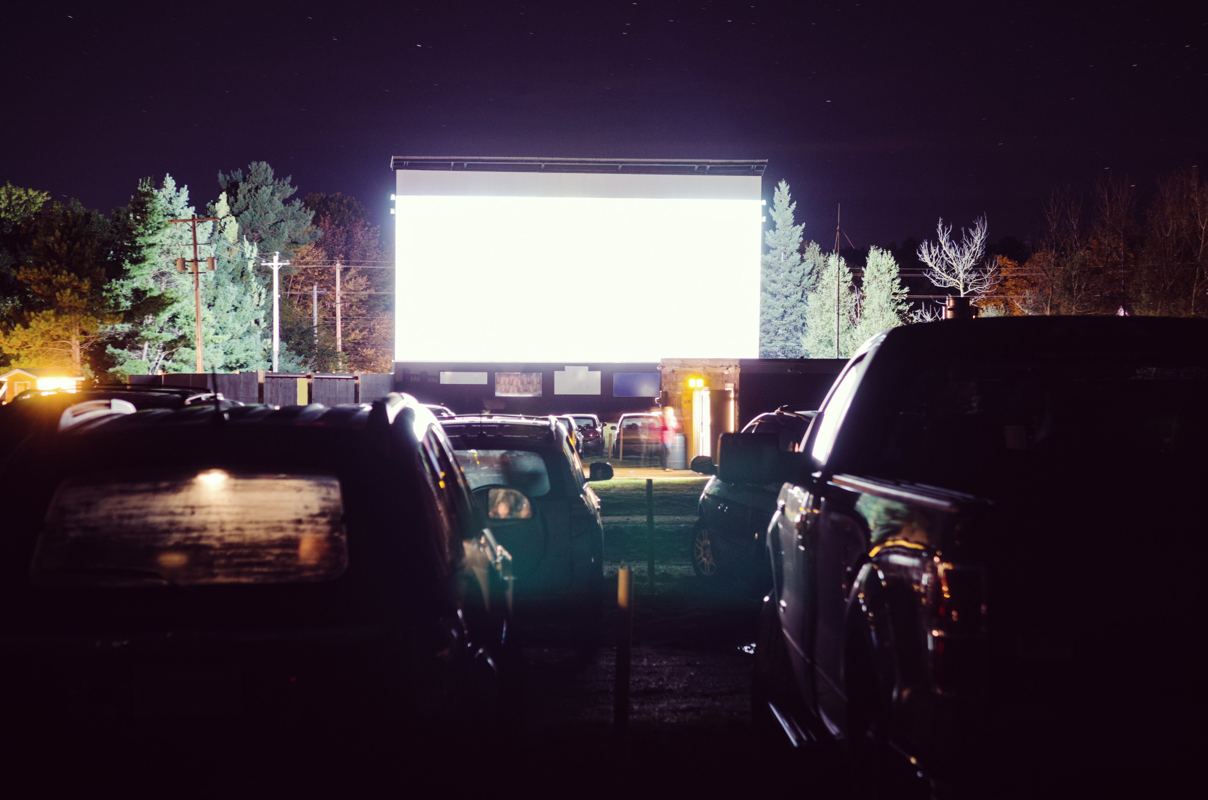 Cars gathered at a drive-in theater with a bright movie screen in the background under a night sky