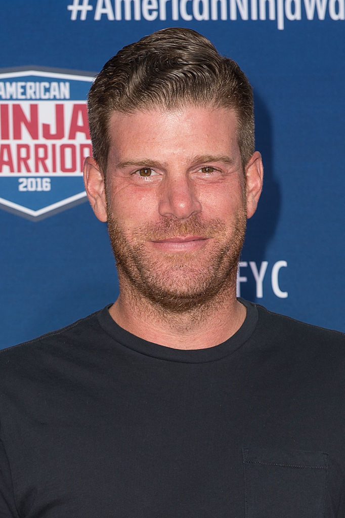 A man with short hair and casual clothing poses in front of an "American Ninja Warrior 2016" backdrop