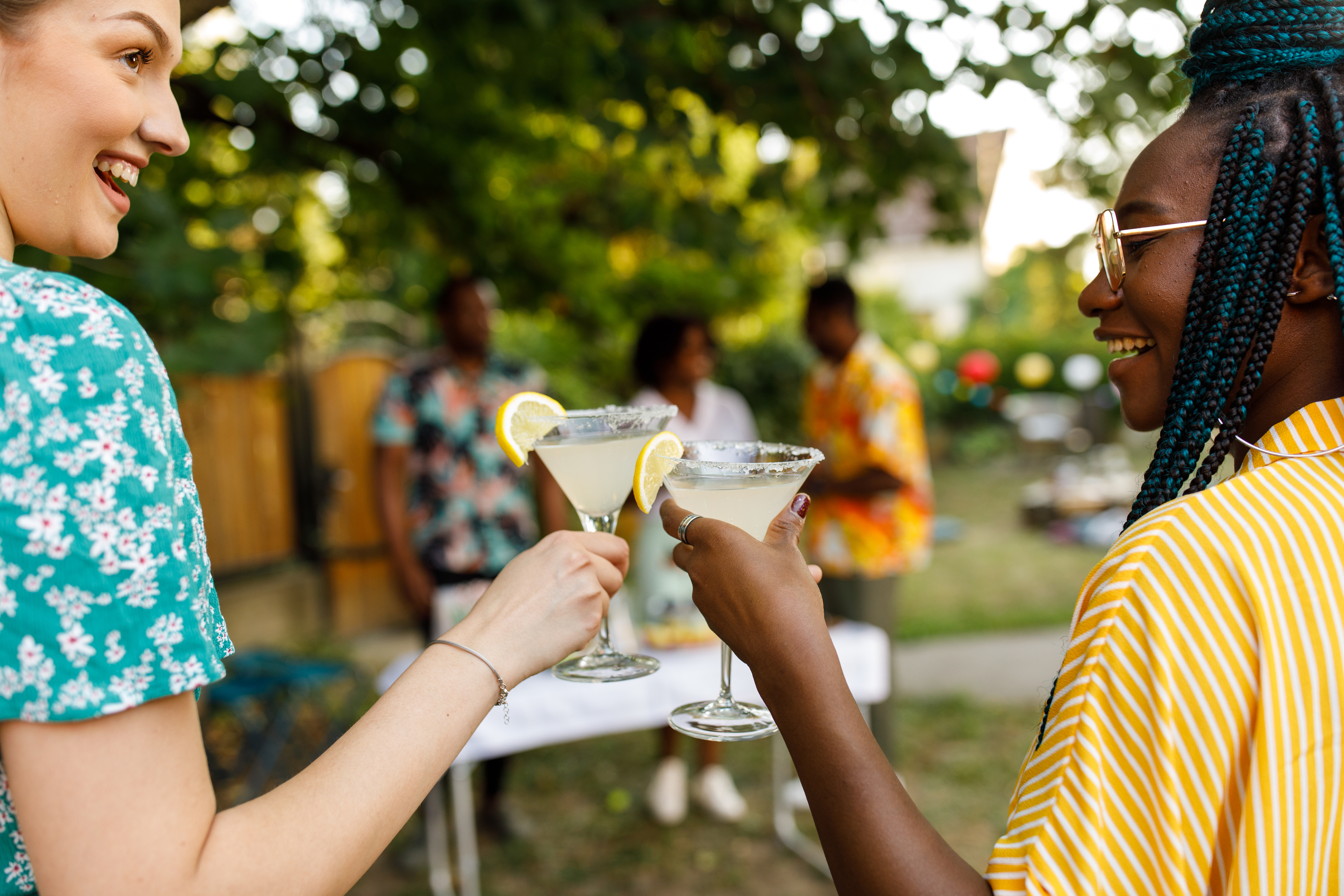 Two women celebrate with cocktails outdoors at a summer gathering, smiling and toasting with glasses garnished with lemon. Other guests are in the background