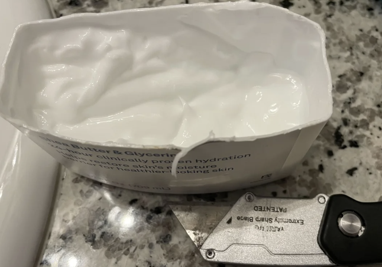 Partially depleted container of cocoa butter and glycerin lotion next to a utility knife