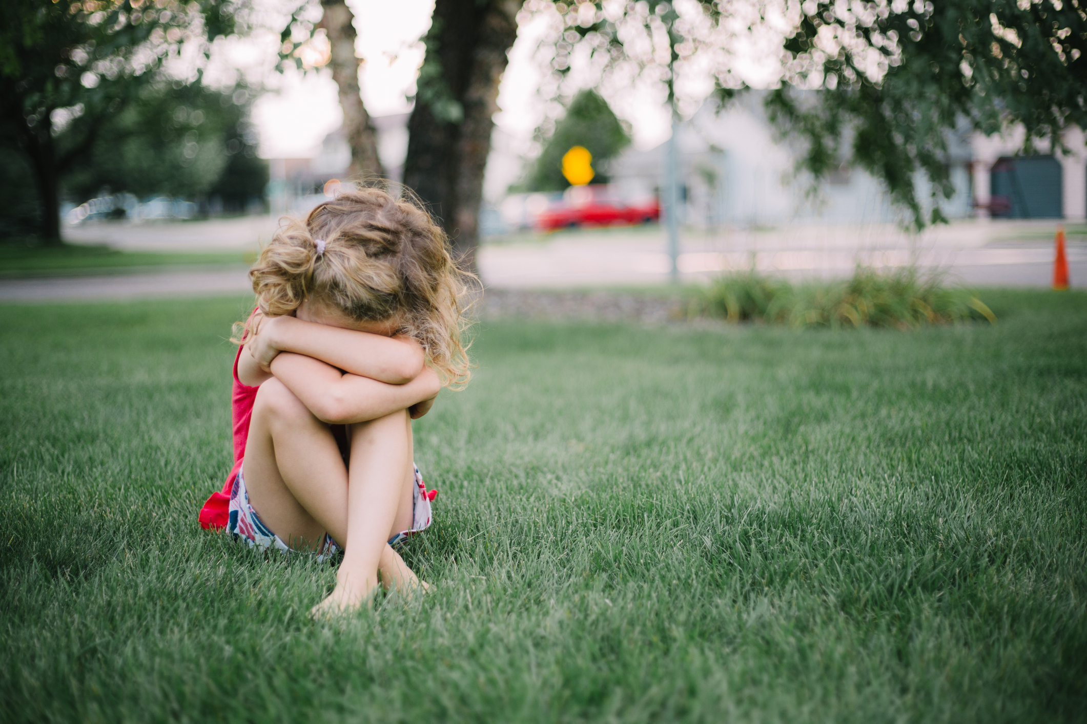 A young child sits on the grass with their arms crossed and head down, appearing upset or deep in thought. Trees and a street are visible in the background