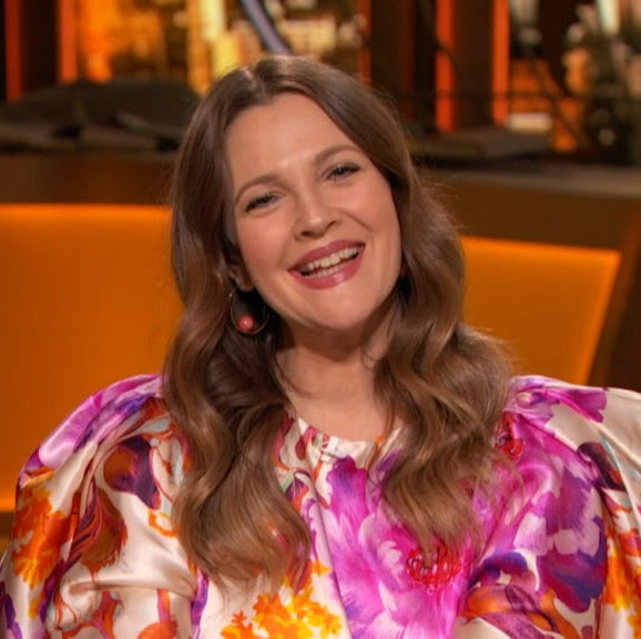 Drew Barrymore is sitting and smiling, dressed in a floral patterned blouse with voluminous sleeves. The background appears to be a cozy, warmly lit indoor setting