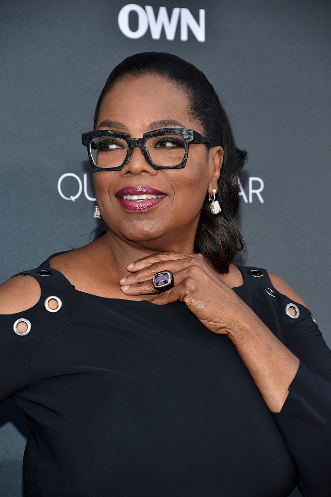 Oprah Winfrey at a red carpet event, wearing a stylish black dress with shoulder cutouts, dark framed glasses, and a large ring, posing with a smile