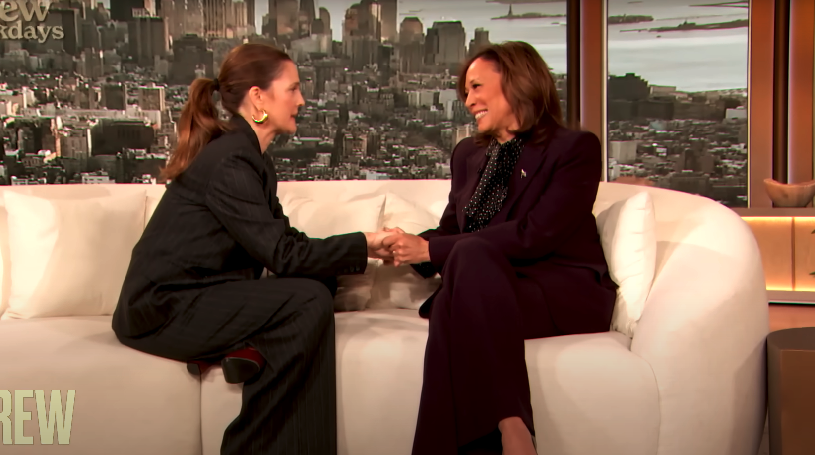 Drew Barrymore, wearing a suit, and Kamala Harris, wearing a suit, sit on a white couch shaking hands and smiling during an interview. A cityscape is in the background