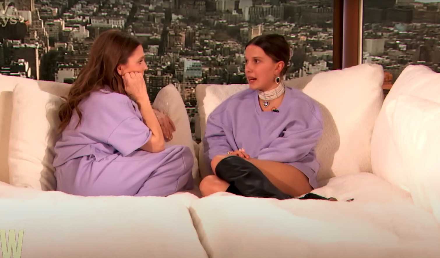 Millie Bobby Brown and Drew Barrymore in conversation on a white couch, wearing loose, light outfits. A cityscape is visible in the background