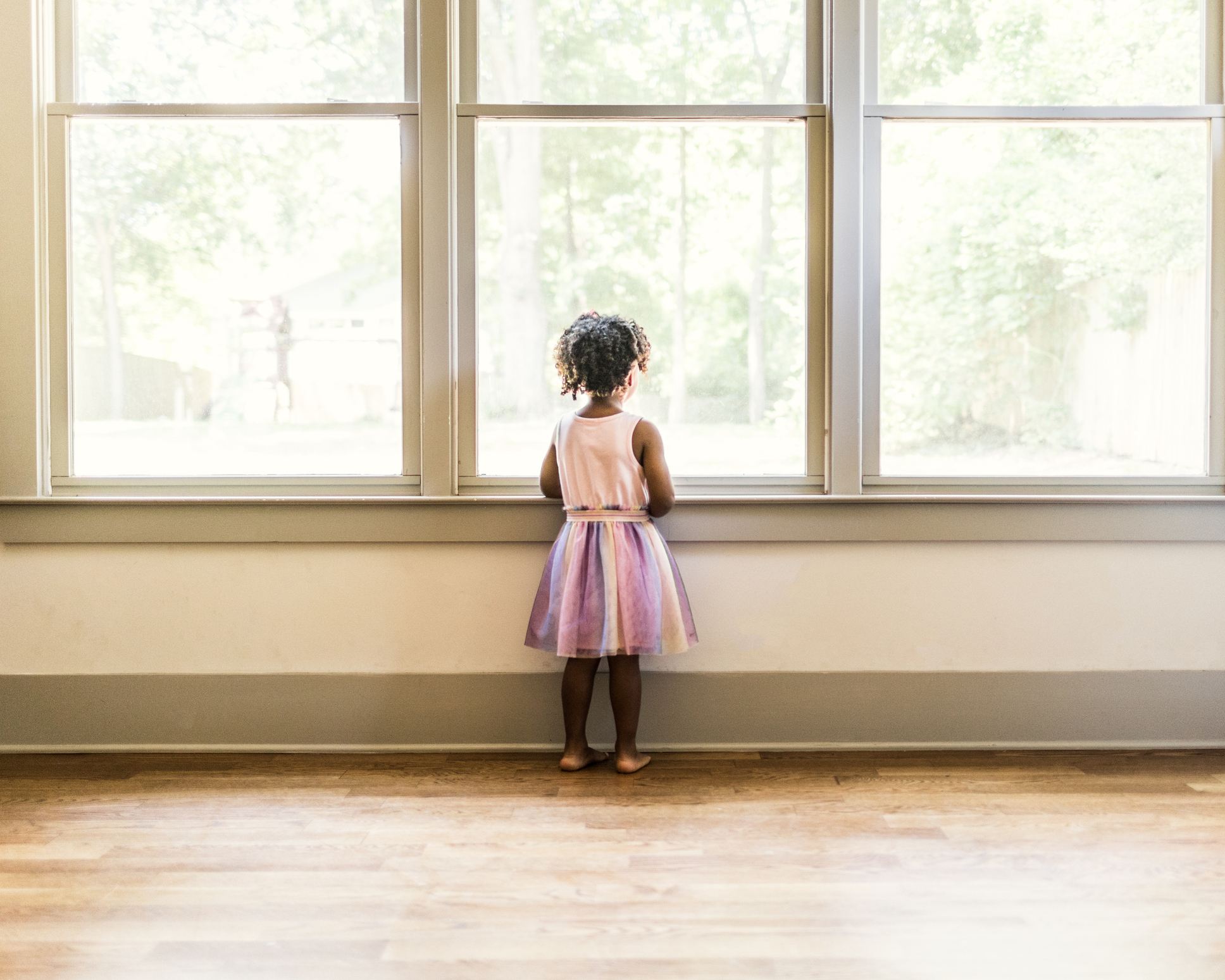 A small child in a dress stands barefoot, looking out of a large window with sunlight streaming in. The child appears to be watching something outside