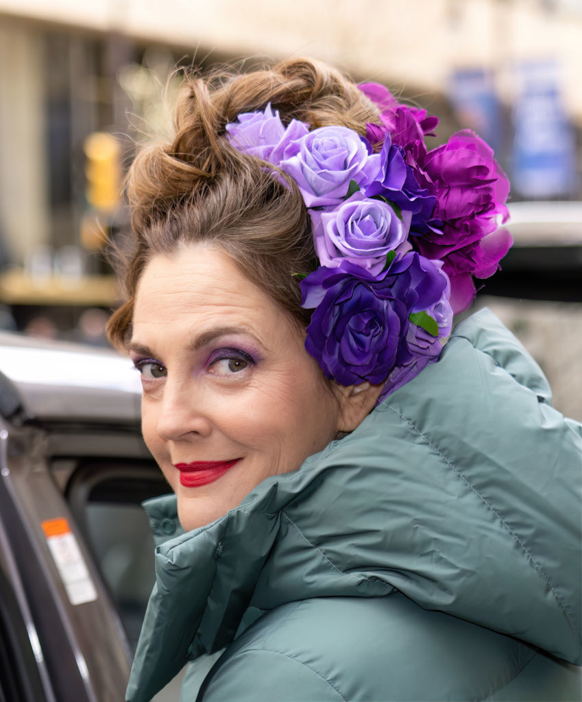Drew Barrymore is wearing a puffer jacket and sporting an elaborate updo adorned with vibrant purple and lavender flowers. She is smiling while looking over her shoulder