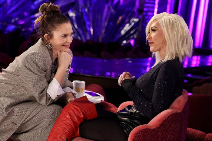 Drew Barrymore and Christina Aguilera sit in plush chairs, engaged in conversation on a stage with bright, decorative backdrop elements