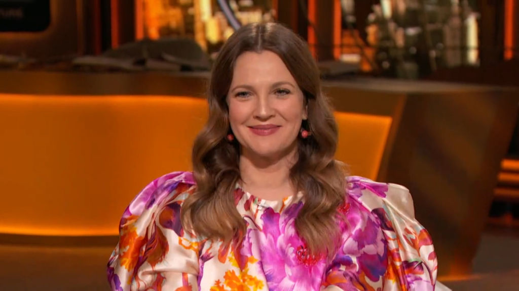 Drew Barrymore is seated in a studio, smiling and wearing a floral-patterned dress with puffy sleeves