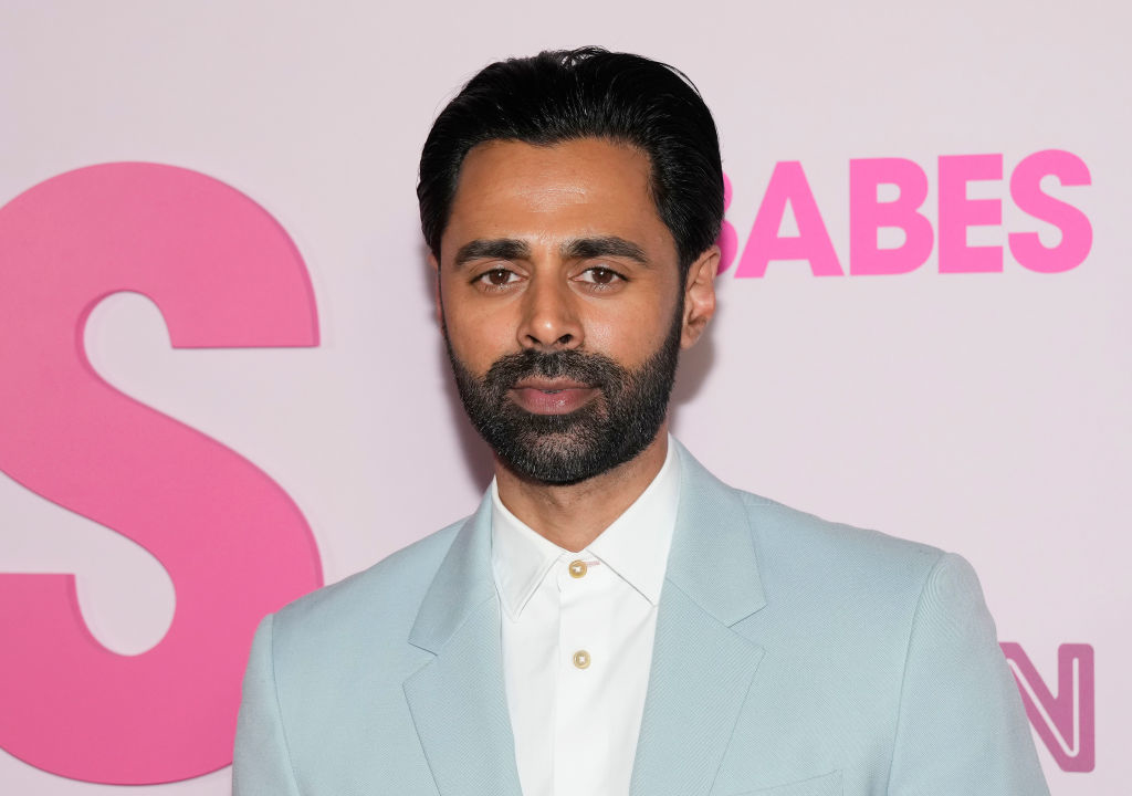Hasan Minhaj at a red carpet event wearing a light-colored suit and white shirt, with a pink background featuring partially visible text