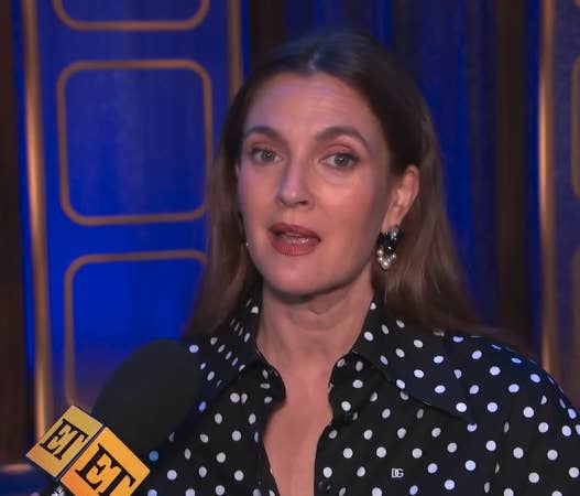 Drew Barrymore speaking into an "Entertainment Tonight" microphone, wearing a black shirt with white polka dots and black earrings