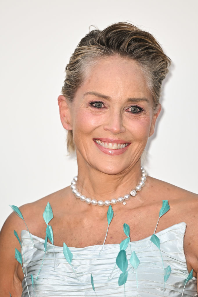 Sharon Stone smiles, wearing a white off-shoulder dress adorned with leaf-like details and a pearl necklace