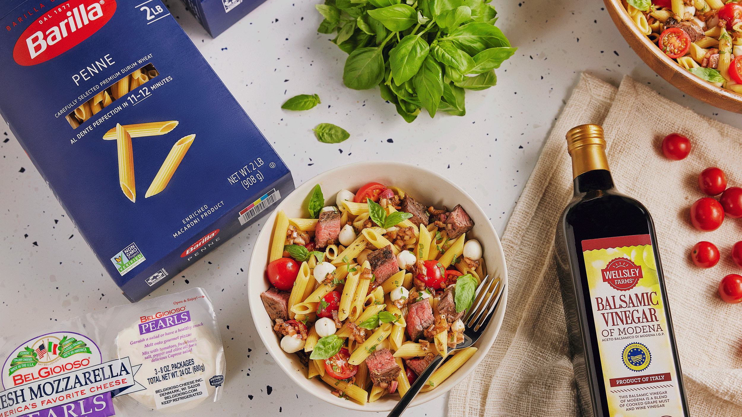 A prepared bowl of penne pasta salad with tomatoes, mozzarella, and greens sits surrounded by pasta boxes, fresh mozzarella, basil, and a bottle of balsamic vinegar