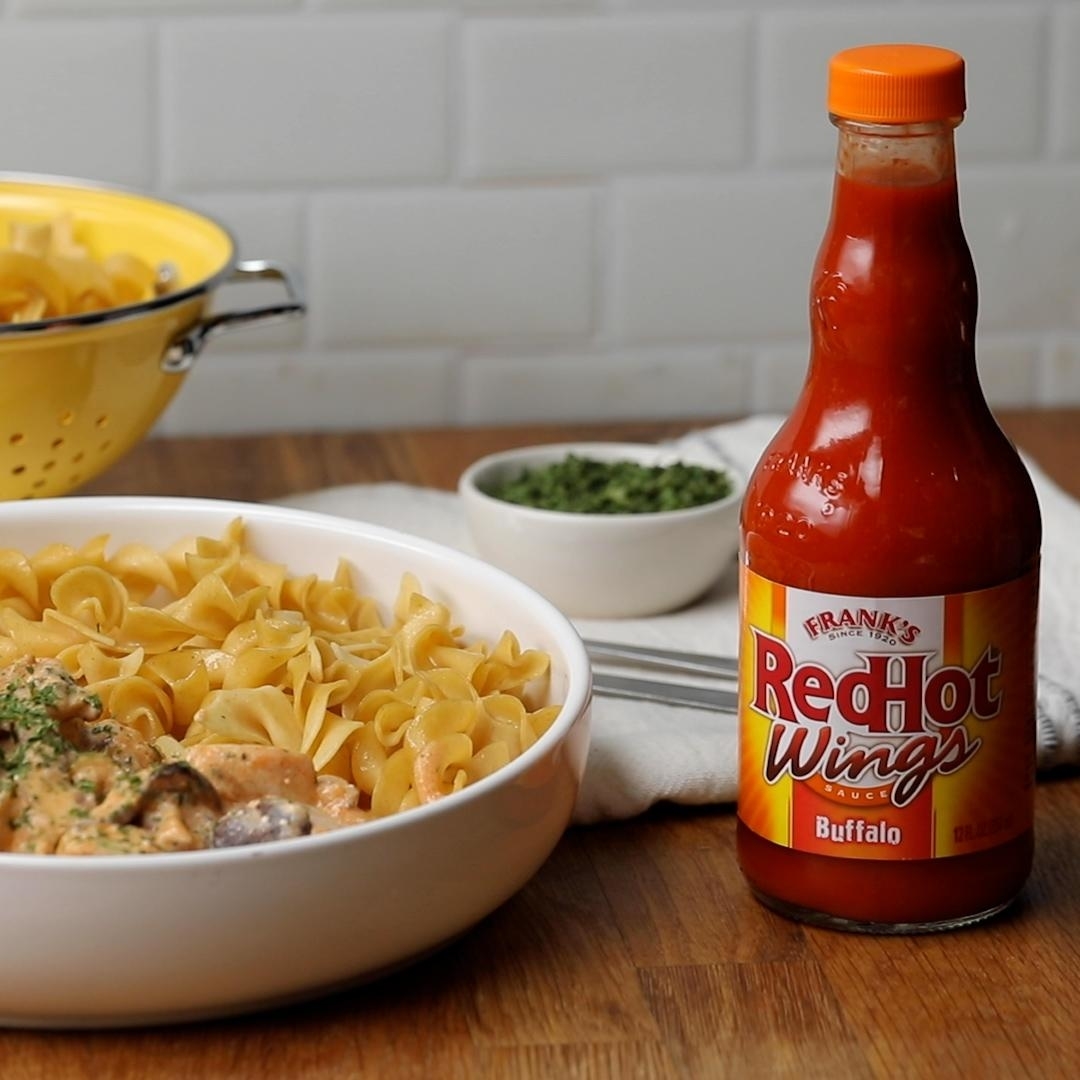 A bowl of pasta with sauce next to a bottle of Frank&#x27;s RedHot Wings Buffalo sauce. There is a colander and a bowl with green herbs in the background