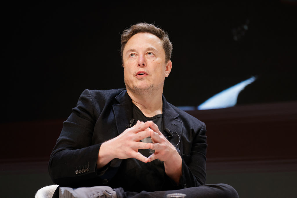 Elon Musk seated, wearing a dark suit, speaking with hands gestured in a talking manner