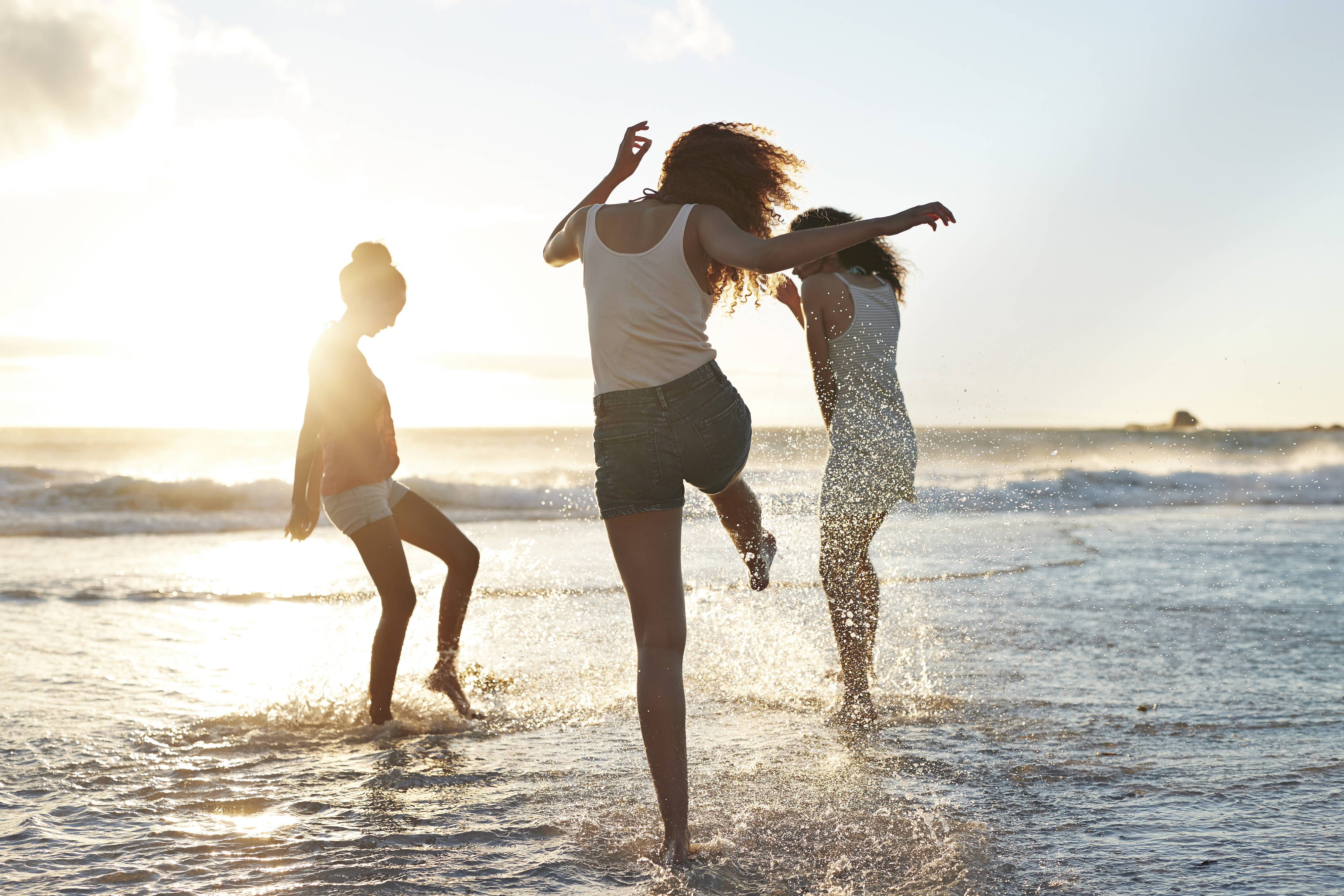 Three people enjoy the beach, splashing in the shallow water at sunset. One person kicks water into the air while the others frolic nearby