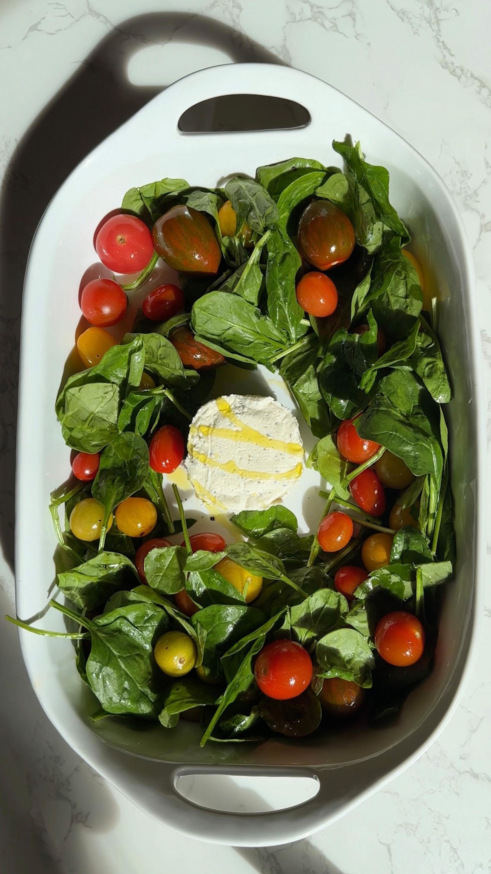 A dish with fresh spinach, cherry tomatoes, and a dollop of creamy cheese in the center, drizzled with olive oil