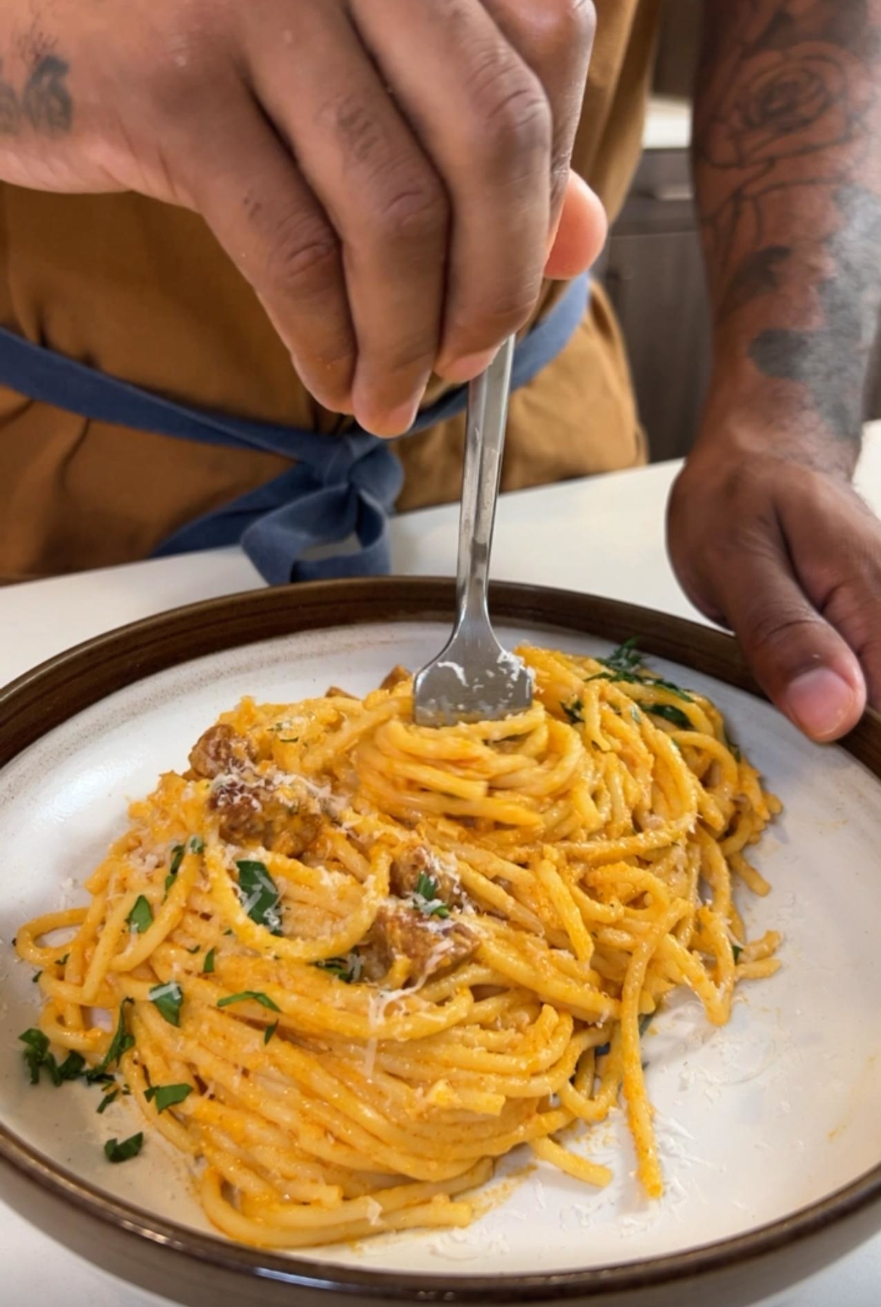 A person with tattoos prepares a plate of creamy pasta with herbs by twirling it with a fork