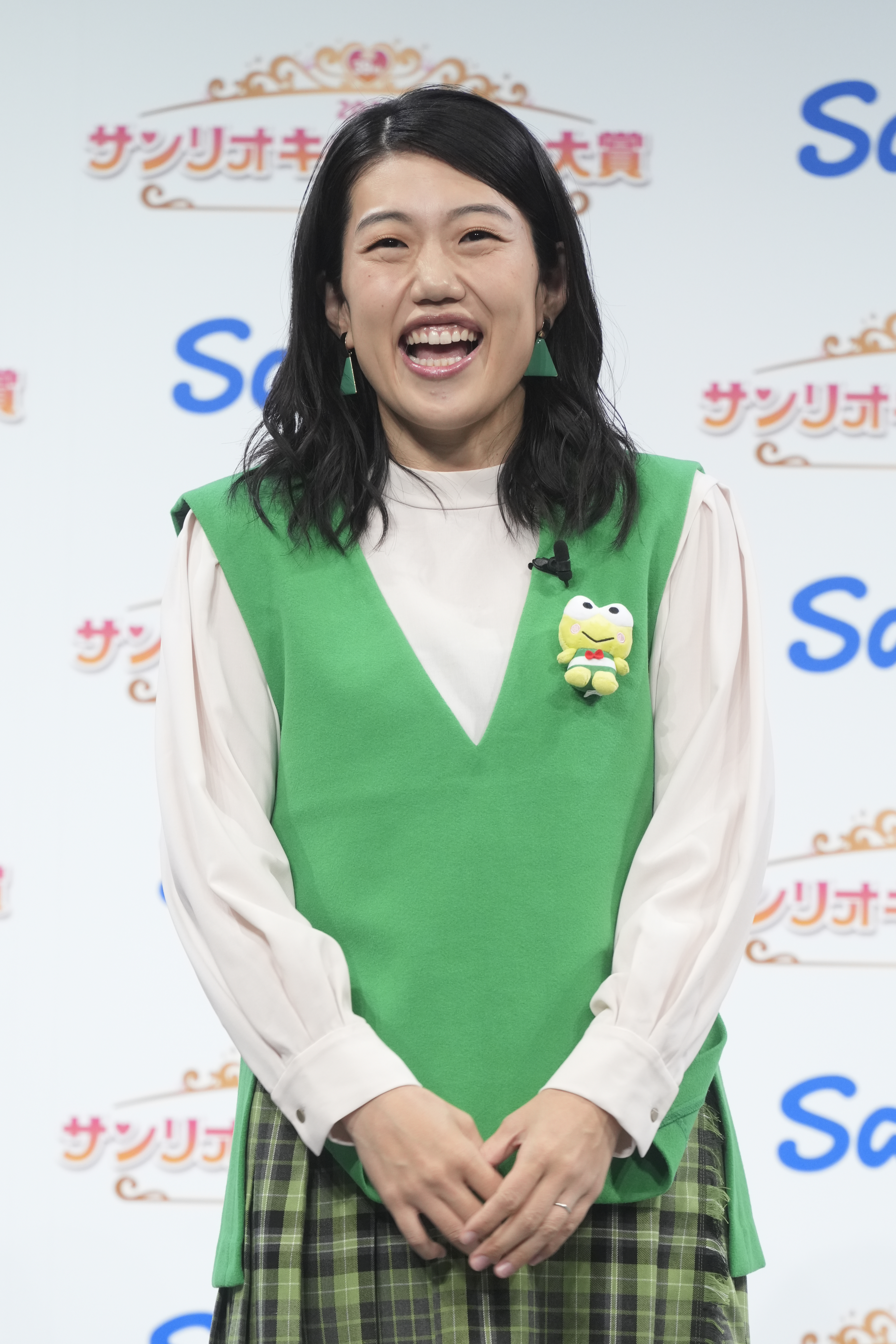 女性が緑色の衣装を着てサンリオキャラクター大賞のイベントに参加しています。胸にカエルのぬいぐるみをつけています。
