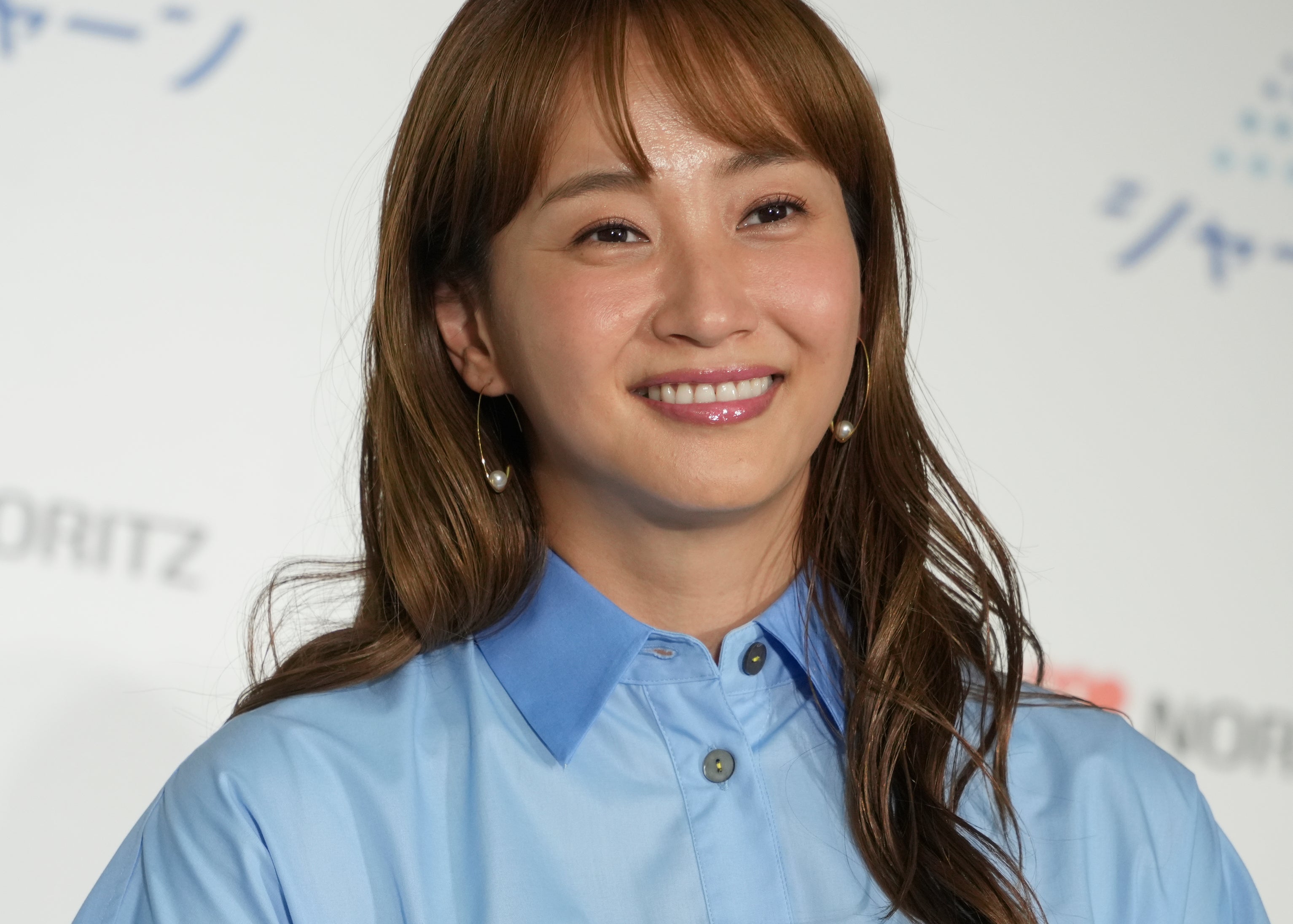 女性がカメラに微笑んでいる姿。青いシャツを着ている。背景に「ノーリツ」やその他の文字が見える。