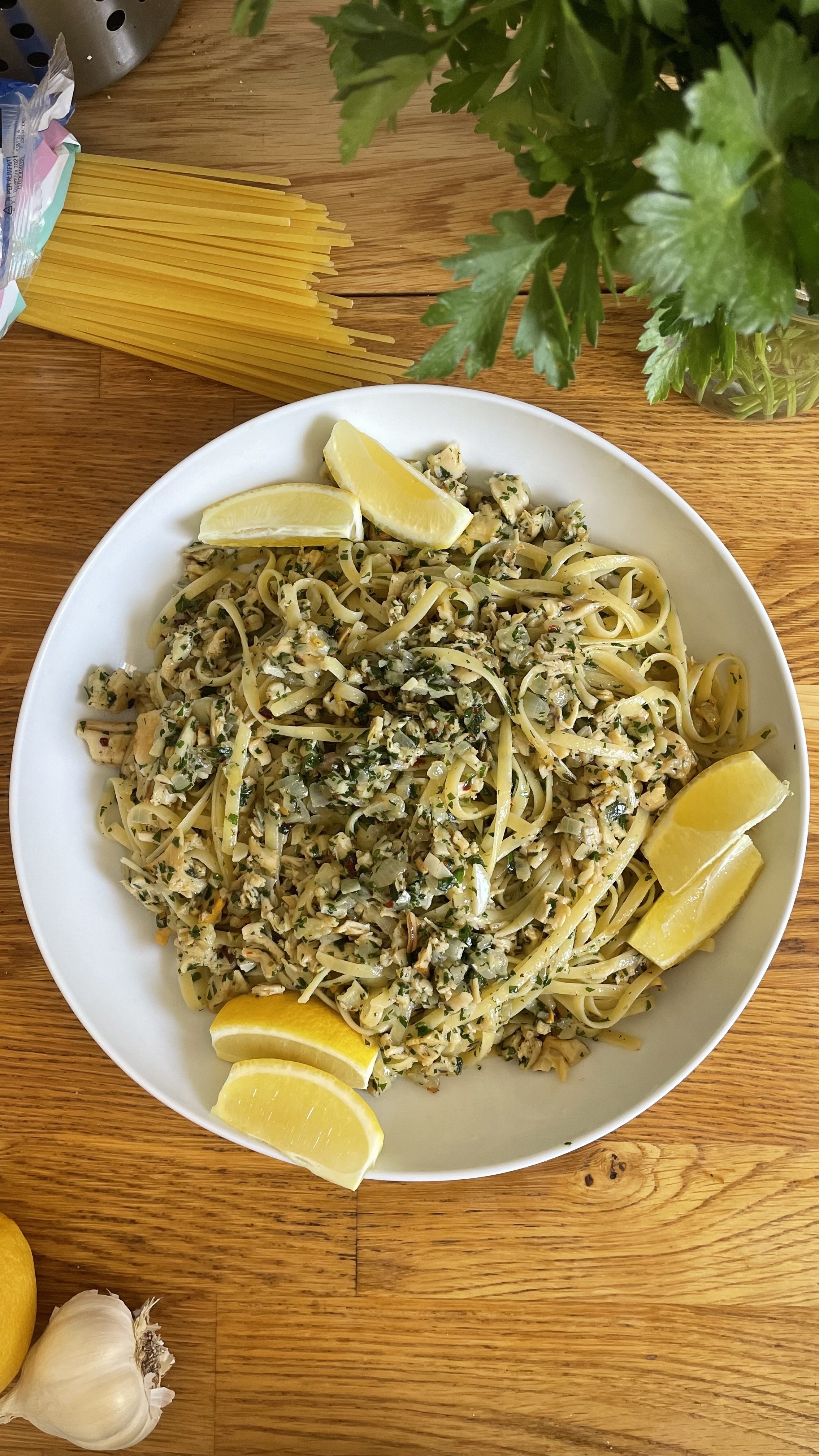 A plate of linguine mixed with herbs and served with three lemon wedges is placed on a wooden table. Uncooked pasta and fresh parsley are also visible