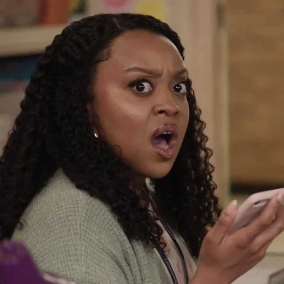 Quinta Brunson looks surprised while holding a smartphone, with her mouth open and eyes wide. She has curly hair and wears a cardigan