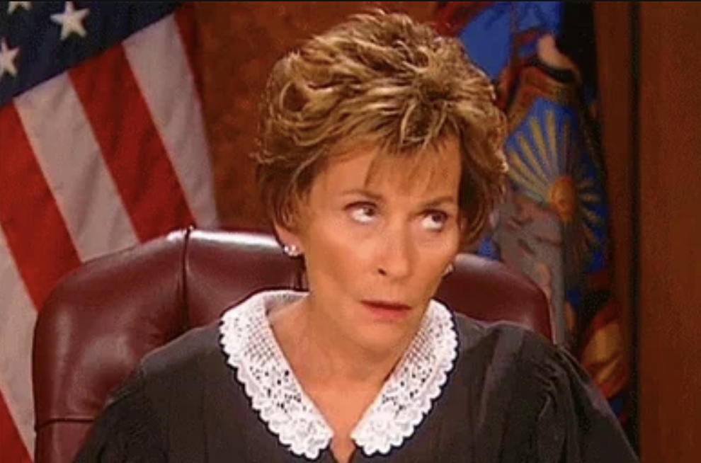 Judge Judy Sheindlin, wearing her traditional judicial robe with a white lace collar, sits at her bench with an expressive look on her face