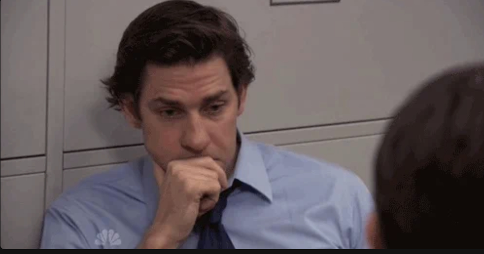 John Krasinski dressed in business attire, looking contemplative with his hand partially covering his mouth, seated in an office setting