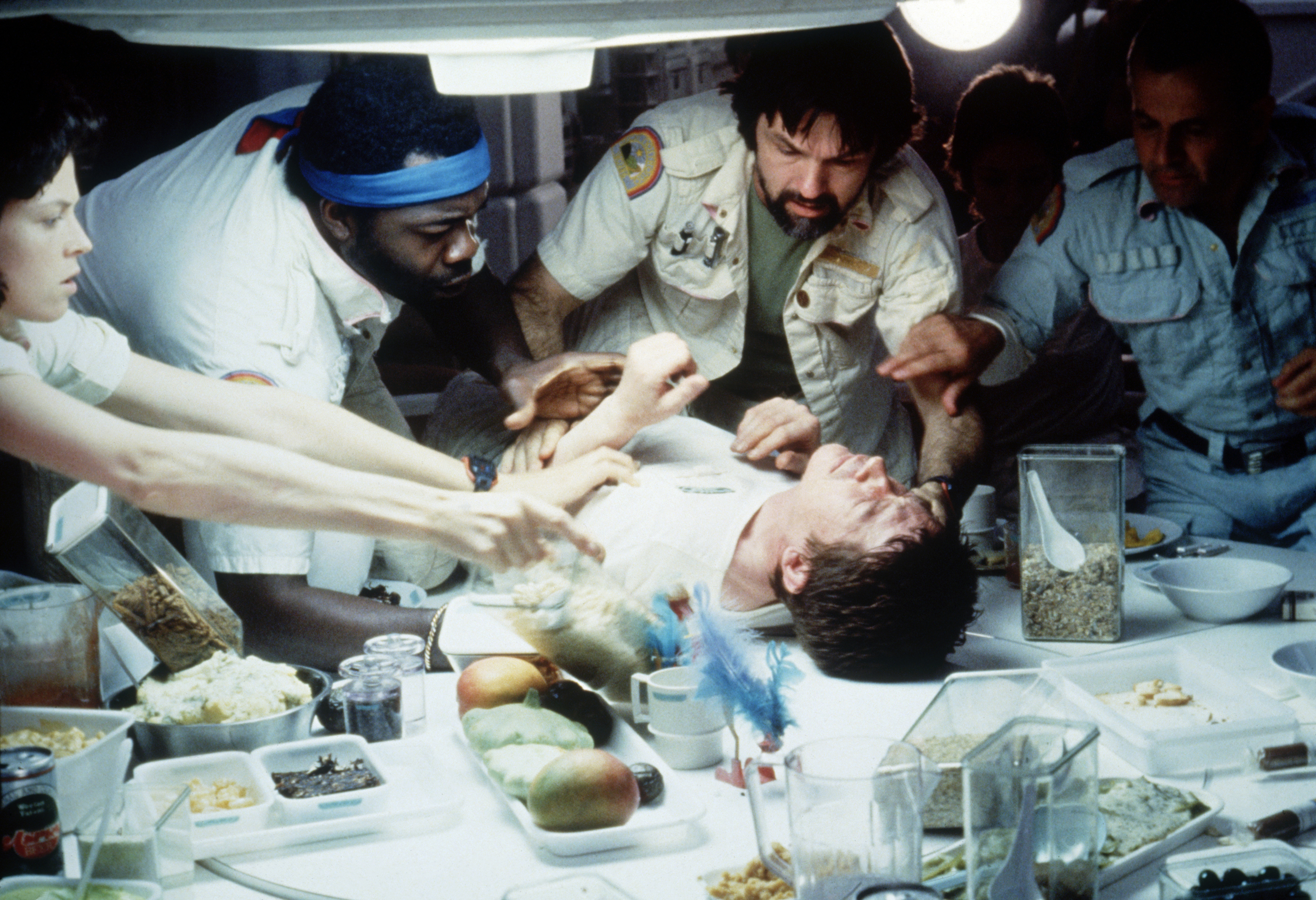 A scene from the movie &quot;Alien&quot; with actors Sigourney Weaver, Yaphet Kotto, Tom Skerritt, and John Hurt. Characters surround John Hurt, who is lying on a table