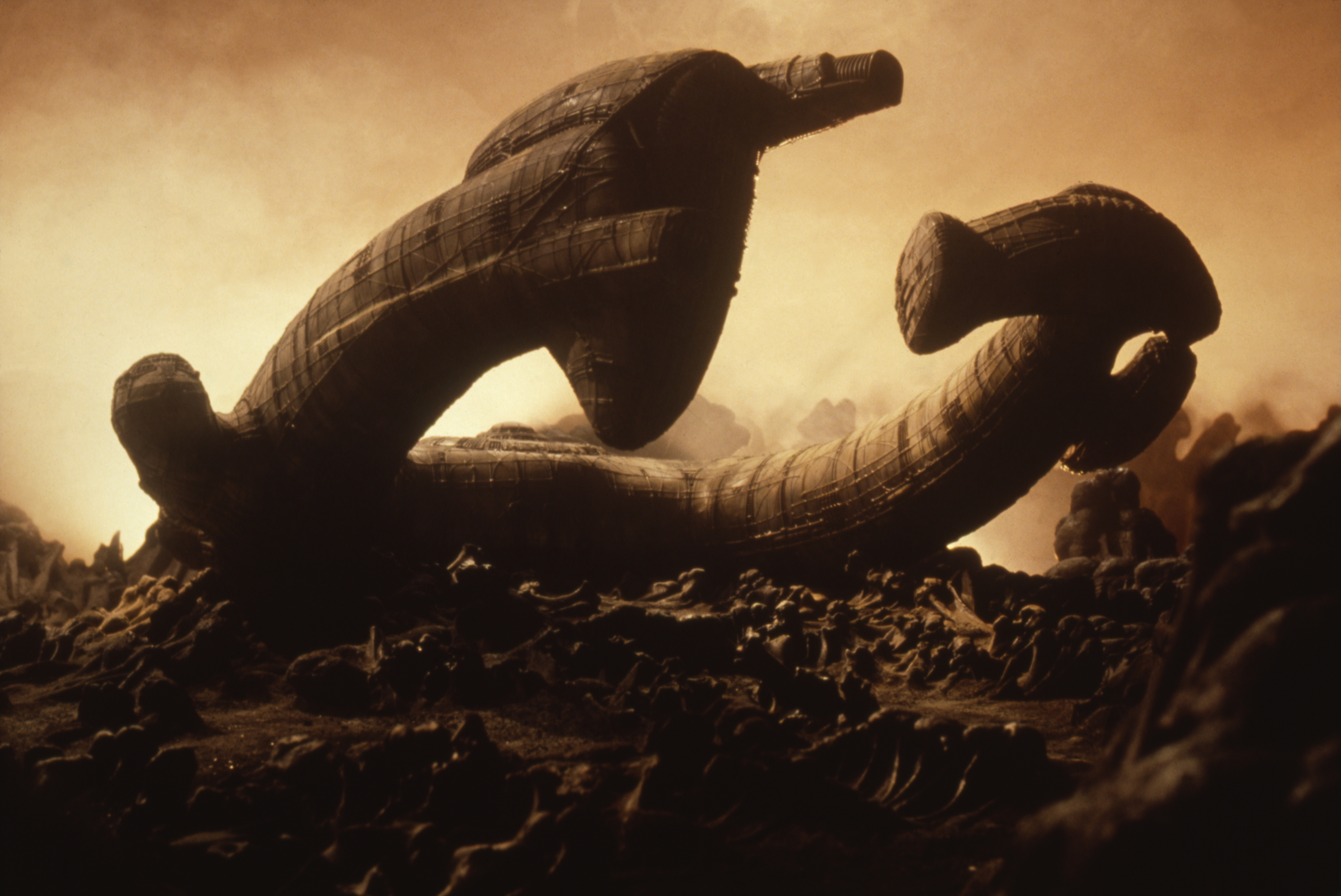 Alien landscape depicting a large, twisted, and decaying spaceship partially buried on a desolate terrain with rocks and mist in the background. No people are visible