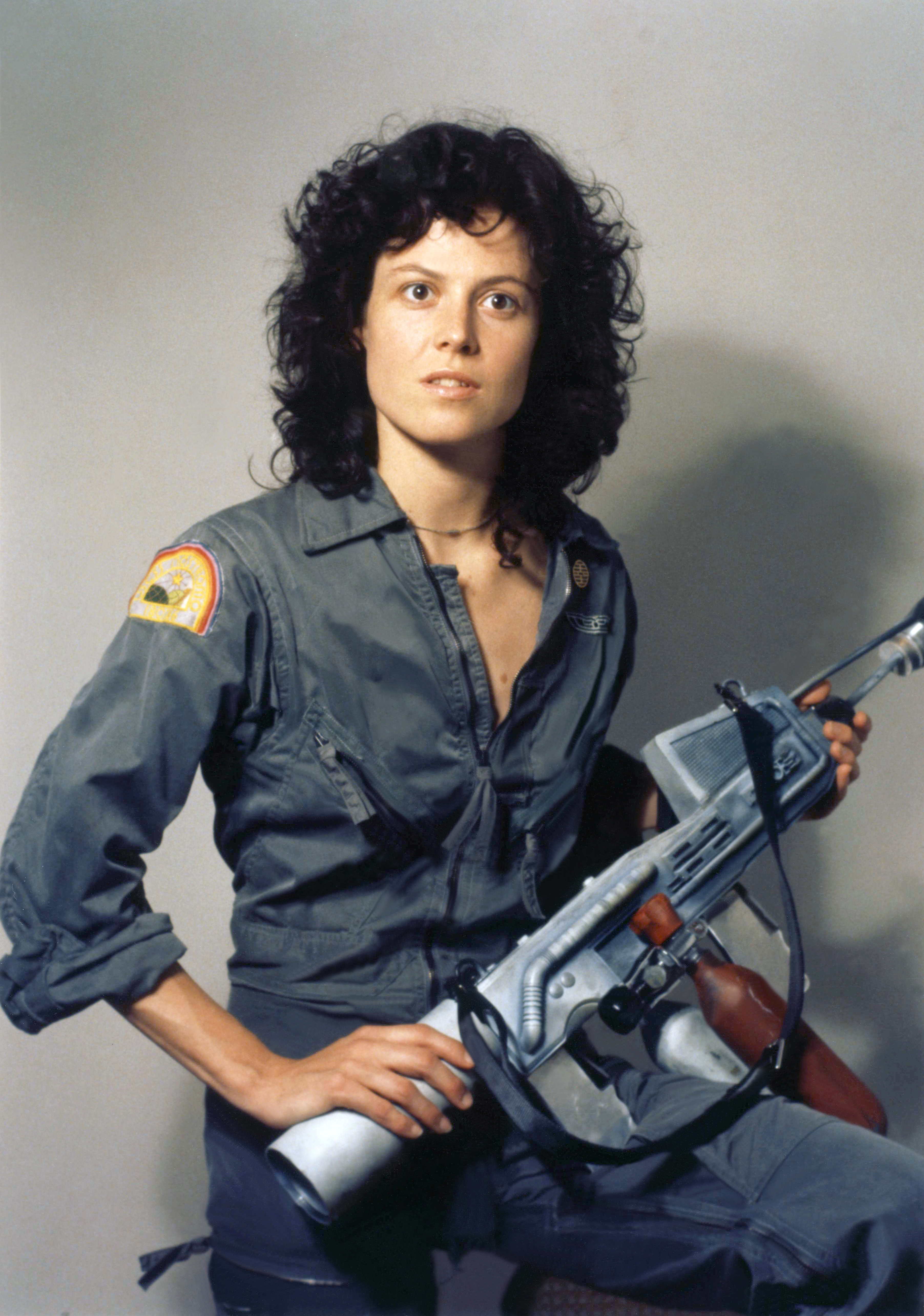 Sigourney Weaver, dressed in a military-style jumpsuit, holds a futuristic weapon in a promotional image for the movie &quot;Alien.&quot;