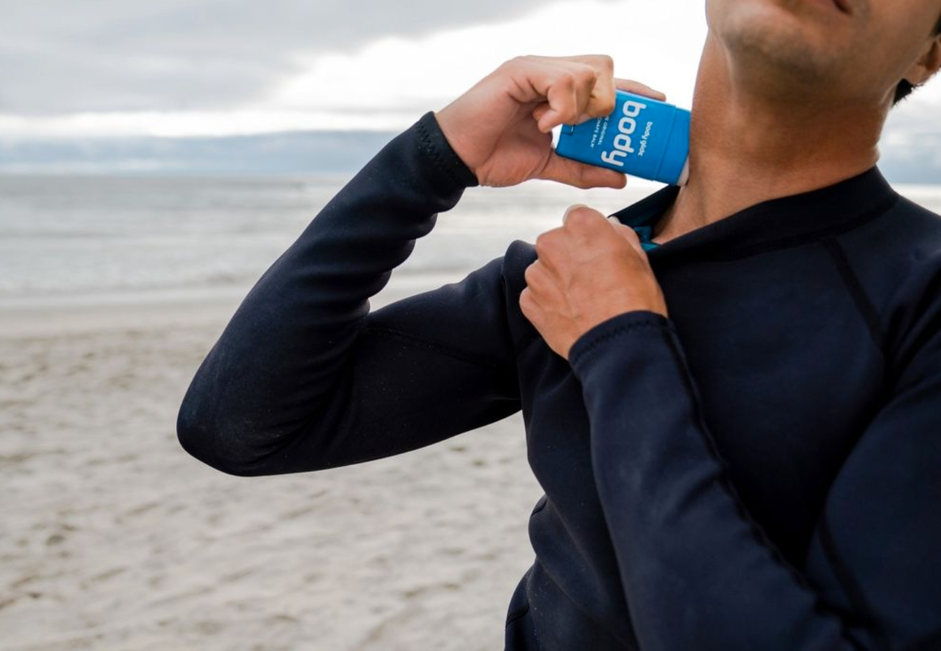 Close-up of a person applying booq sunscreen to their neck at the beach while wearing a long-sleeved shirt