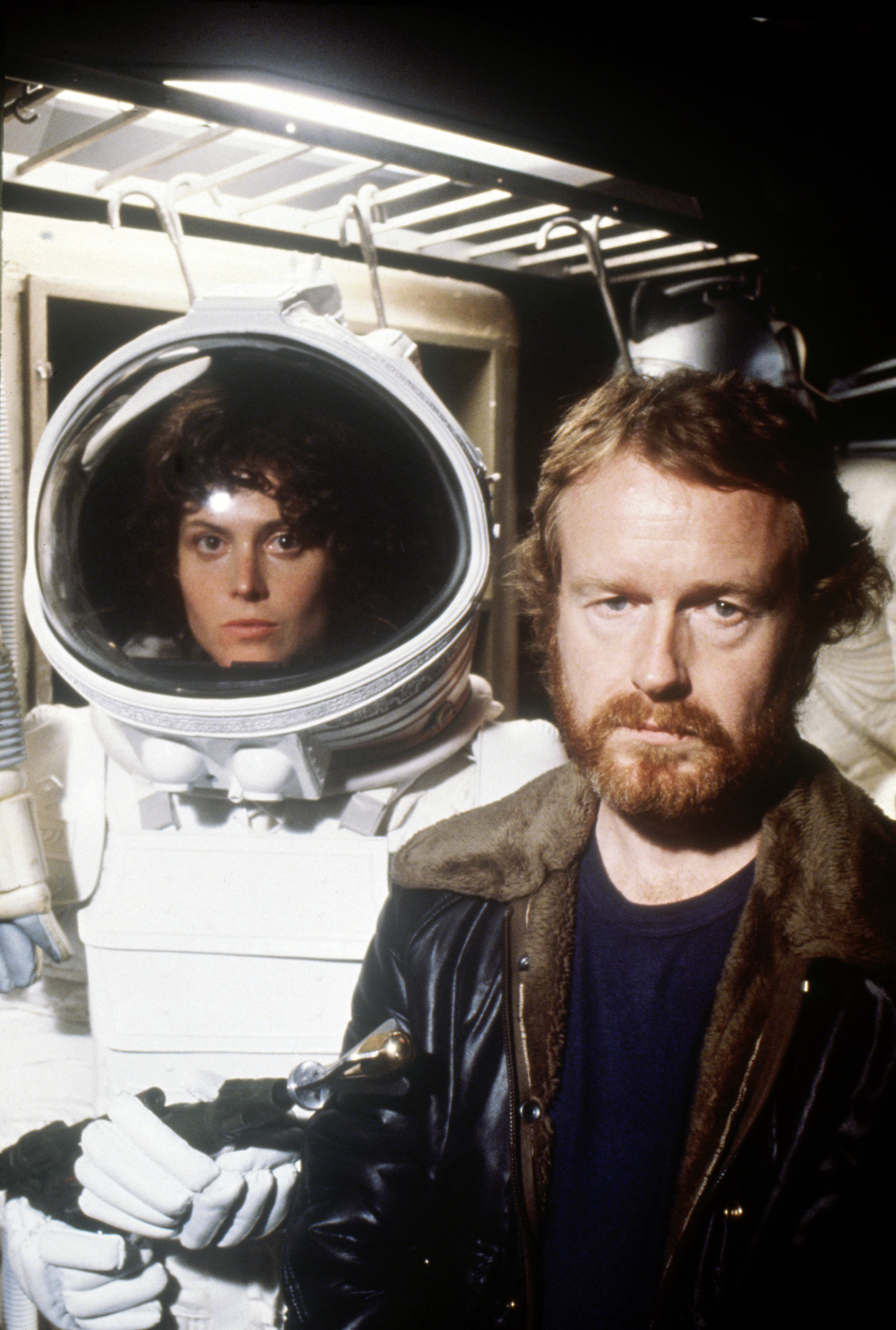 Sigourney Weaver, in a space suit, stands next to Ridley Scott. Both appear serious, with a sci-fi film set in the background